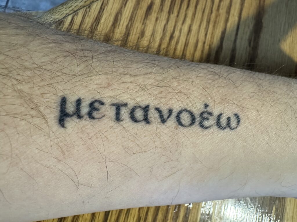 A close-up of a person's forearm featuring a tattoo of the Greek word 'μετάνοια,' which means 'change of mind,' symbolizing personal growth and leadership.